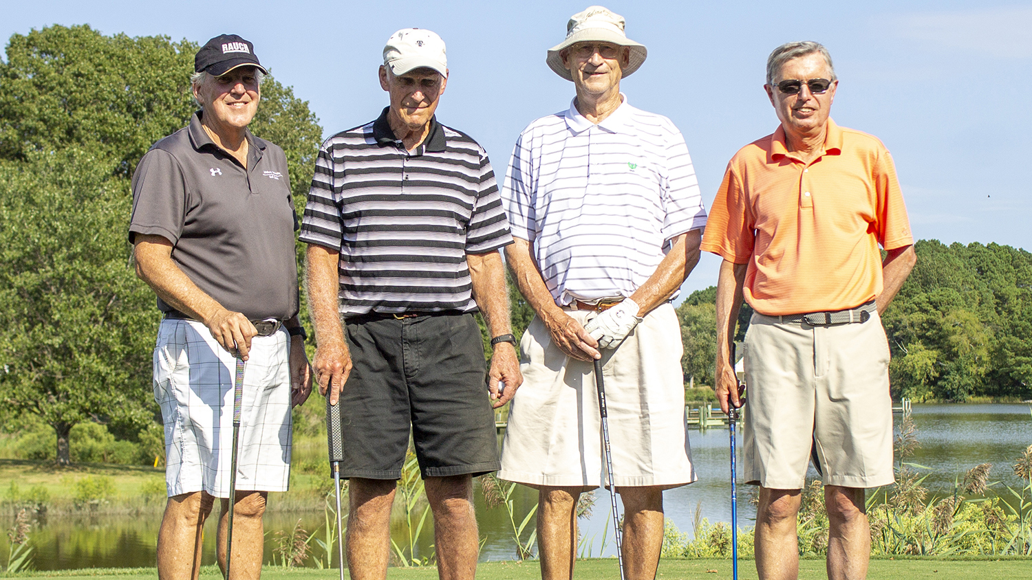 Four men stand in front of a lake, holding golf clubs, on a golf course. It is sunny and they are dressed for summer.