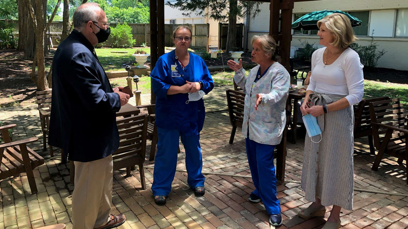 Three women and a man stand around talking. One woman is talking and gesturing with her hands. Two of the women are dressed in scrubs.