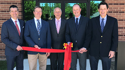Five gentlemen stand in front of a building. They are all holding a red ribbon, which one of them is about to cut.
